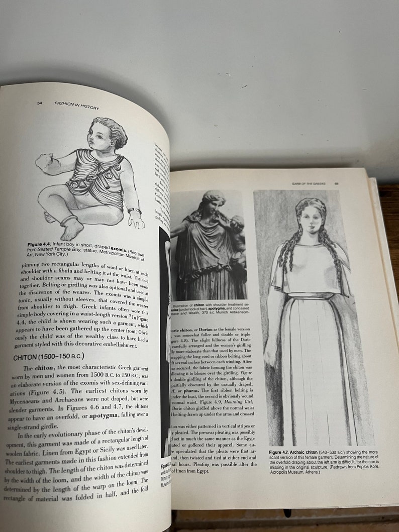 May include: A black and white illustration of a woman wearing a long, flowing garment called a chiton. The chiton is belted at the waist and has a draped overfold. The illustration is from a book about fashion history.