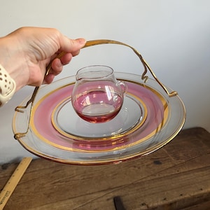 May include: A vintage glass serving tray with a pink and gold rim, and a small glass creamer pitcher. The tray has a gold metal handle. The tray is held by a person.