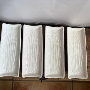 May include: A set of four rectangular white ceramic serving dishes, each featuring a raised, embossed corn cob design in the center. The dishes have a smooth, glossy finish and a decorative rope-style border along the edges of the rectangular form.