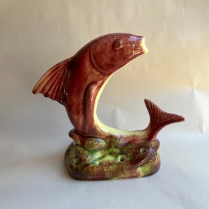 May include: A ceramic fish figurine with a red and green glaze. The fish is jumping out of the water and has a small fish tail behind it. The base of the figurine is decorated with green and brown leaves.