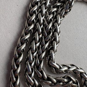 Pu&ograve; includere: Primo piano di una catena color argento con un motivo a spiga intrecciata. La catena &egrave; composta da maglie interconnesse, creando una superficie strutturata. Il metallo ha una qualit&agrave; lucida e riflettente, su sfondo grigio chiaro.