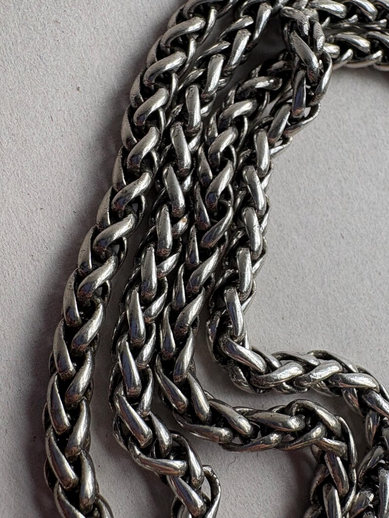 Pu&ograve; includere: Primo piano di una catena color argento con un motivo a spiga intrecciata. La catena &egrave; composta da maglie interconnesse, creando una superficie strutturata. Il metallo ha una qualit&agrave; lucida e riflettente, su sfondo grigio chiaro.