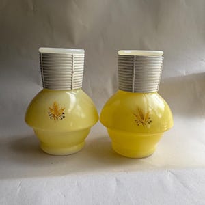May include: Two matching yellow glass shakers with a wide, rounded base and a narrow, ribbed white plastic neck. Each shaker features a small, centered graphic of a golden wheat stalk. The tops are finished with a flat, white plastic lid.