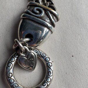 Pu&ograve; includere: Primo piano di una chiusura e di un ciondolo per collana argentati. La chiusura ha un design a spirale decorativo. Un ciondolo a forma di cuore con le iniziali "MB" pende da un anello decorativo. La catena &egrave; in stile intrecciato.