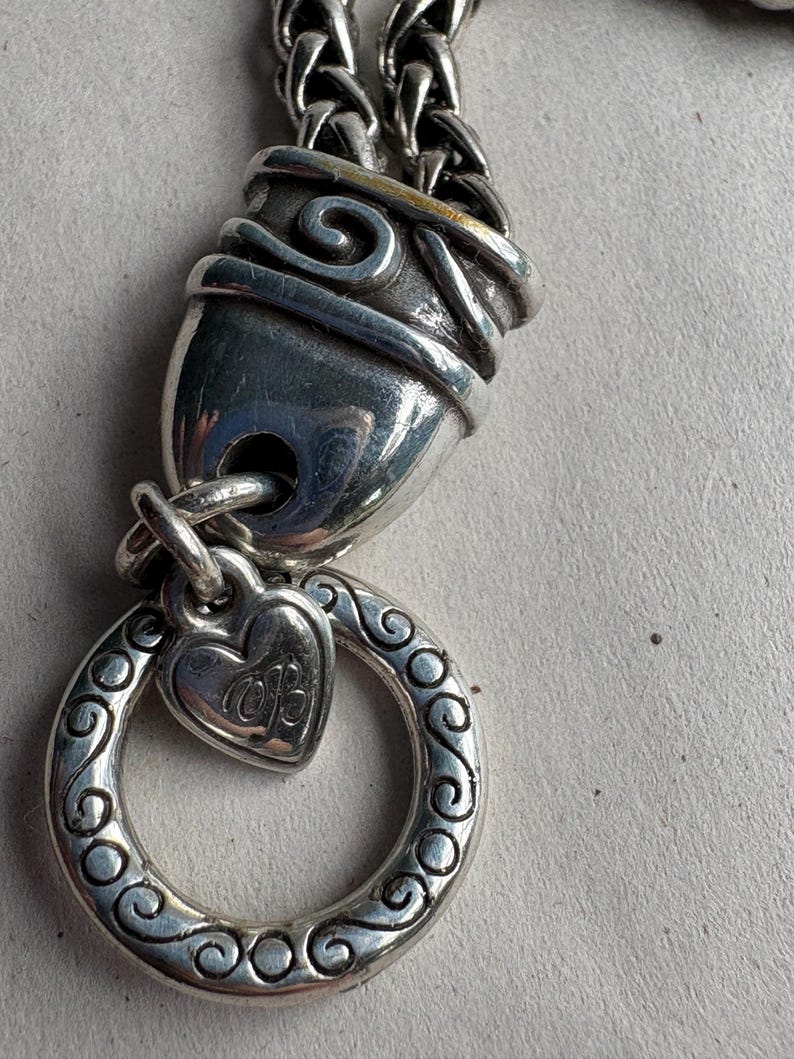 Pu&ograve; includere: Primo piano di una chiusura e di un ciondolo per collana argentati. La chiusura ha un design a spirale decorativo. Un ciondolo a forma di cuore con le iniziali "MB" pende da un anello decorativo. La catena &egrave; in stile intrecciato.