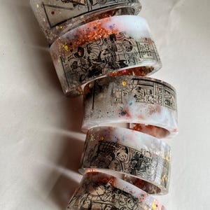 May include: Stack of clear resin bracelets with black and white illustrations, copper flakes, and glitter. The bracelets have a wide cuff design. The illustrations appear to be of people and scenes.