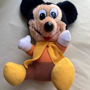 Op de afbeelding: Een pluche Mickey Mouse-speelgoed met zwarte oren, een bruin gezicht en grote ogen met witte pupillen. Het speelgoed draagt een oranje outfit met een gele sjaal en gele voeten. Het speelgoed zit met de armen omhoog.