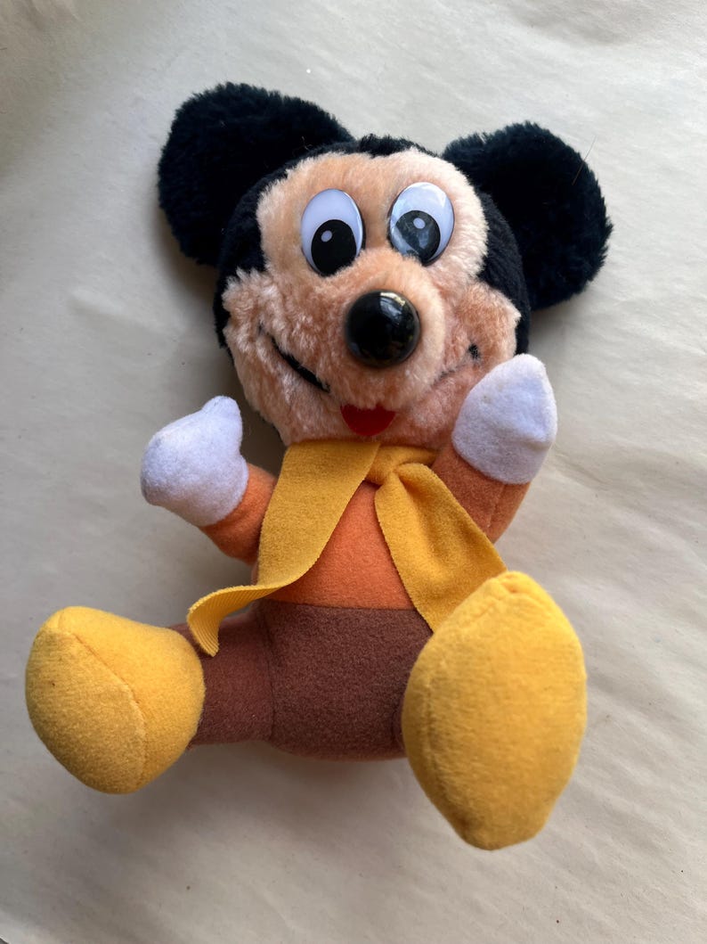 Op de afbeelding: Een pluche Mickey Mouse-speelgoed met zwarte oren, een bruin gezicht en grote ogen met witte pupillen. Het speelgoed draagt een oranje outfit met een gele sjaal en gele voeten. Het speelgoed zit met de armen omhoog.