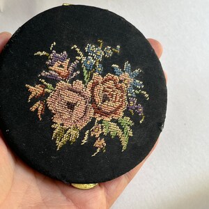 May include: A round black compact with a floral design embroidered on the top. The design features pink and purple roses with green leaves and blue forget-me-nots.