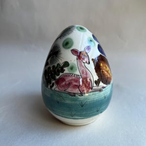 May include: A ceramic egg-shaped decorative item with a hand-painted design. The design features a resting deer in shades of pink and brown, surrounded by green foliage and blue accents. The base is a teal color.
