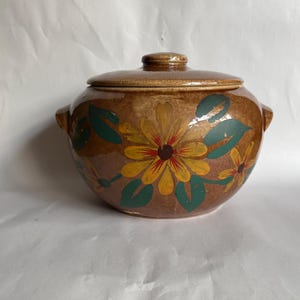 May include: A round brown ceramic casserole dish with a matching lid and two side handles. The exterior features a hand-painted floral pattern with large yellow flowers, dark red centers, and dark green leaves against a mottled brown glaze.