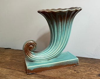 May include: A turquoise and brown ceramic vase shaped like a horn with a flared opening. The vase is sitting on a wooden surface.