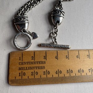 Pu&ograve; includere: Collana color argento con chiusura e catena decorative. La collana presenta una chiusura a toggle e una chiusura ad anello. Un righello di legno con segni in centimetri e millimetri &egrave; posto accanto alla collana.