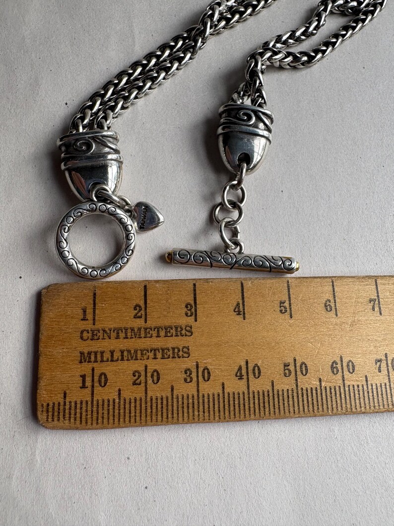 Pu&ograve; includere: Collana color argento con chiusura e catena decorative. La collana presenta una chiusura a toggle e una chiusura ad anello. Un righello di legno con segni in centimetri e millimetri &egrave; posto accanto alla collana.