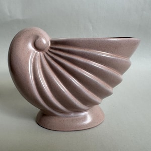 May include: A dusty rose ceramic vase shaped like a nautilus shell. The vessel features a spiraled design with raised, radiating ridges that extend from the center curl to the wide, open top. It stands on a circular base with a smooth finish.