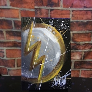 Może przedstawiać: Mały obraz na płótnie z czarnym tłem i żółtym symbolem błyskawicy w białym kółku. Błyskawica jest obwiedziona złotem, a koło ma złotą obwódkę. Obraz ma efekt rozprysku białej i złotej farby.