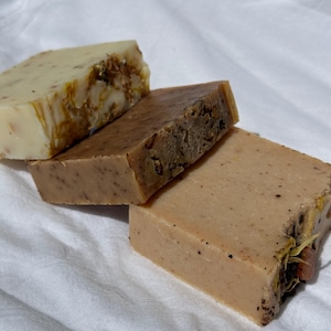 May include: Four handmade soap bars in varying shades of beige and brown, arranged on a white cloth. The soaps have visible natural ingredients like dried flowers and seeds. The bars are rectangular and appear to be freshly made.