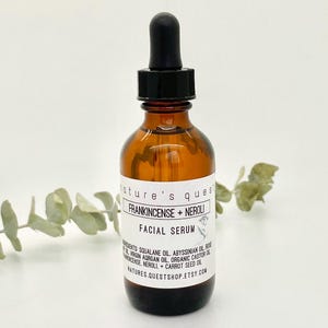 Peut inclure: Un flacon en verre brun avec un compte-gouttes noir, &eacute;tiquet&eacute; "Nature's Quest Frankincense + Neroli Facial Serum". Le flacon est pos&eacute; sur une surface blanche avec une branche verte feuillue en arri&egrave;re-plan.
