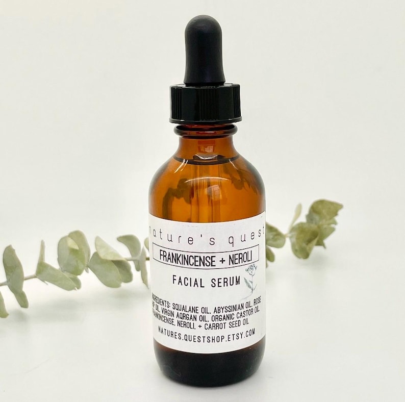 Peut inclure: Un flacon en verre brun avec un compte-gouttes noir, &eacute;tiquet&eacute; "Nature's Quest Frankincense + Neroli Facial Serum". Le flacon est pos&eacute; sur une surface blanche avec une branche verte feuillue en arri&egrave;re-plan.
