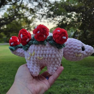 May include: A crocheted, light pink dinosaur-like creature with red mushroom accents. The mushrooms have white spots and green leaves. The creature has a small yellow flower design on its side and a black eye.