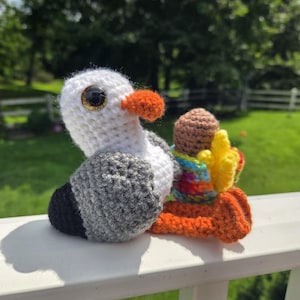 May include: A handmade crochet seagull with a white head, gray wings, and a black tail. It has an orange beak and a large, gold-rimmed eye. The seagull is perched on an orange and rainbow-colored base.