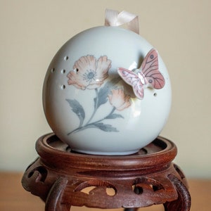 Op de afbeelding: Een witte porseleinen ornament met een bloemenmotief en een roze vlinder. De ornament staat op een donkerbruine houten standaard met ingewikkelde houtsnijwerk.