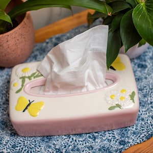 May include: A pink ceramic tissue box holder with a white interior and a floral design featuring yellow butterflies and white daisies. The tissue box is sitting on a blue and white patterned table runner.