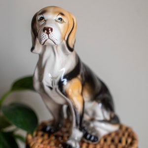 May include: A ceramic figurine of a beagle dog sitting on a wicker stool. The dog is painted in shades of brown, black, and white.