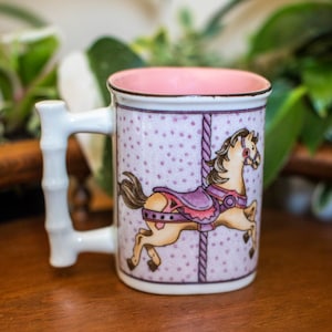 Op de afbeelding: Een witte keramische mok met een roze binnenkant en een bamboe handvat. De mok heeft een kleurrijke illustratie van een draaimolenpaard met paarse en roze accenten op een lavendelkleurige achtergrond met witte stippen.