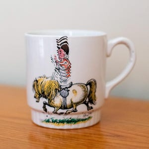 May include: White ceramic mug featuring a cartoon illustration of a person riding a horse. The person is wearing a hat and a striped shirt. The horse is brown and white.