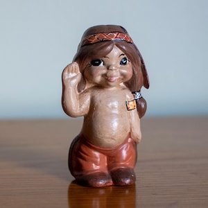 以下が含まれることがあります： オレンジと白の幾何学模様が施された茶色のヘッドバンドをつけたネイティブアメリカンの子供の小さなセラミック製フィギュア。フィギュアは茶色、オレンジ、白の色合いで塗られており、友好的な表情をしています。