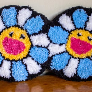 Puede incluir: Dos decoraciones de alfombra de flores sonrientes, azules y blancas, con contornos negros. Las flores tienen un centro amarillo con una boca rosa.