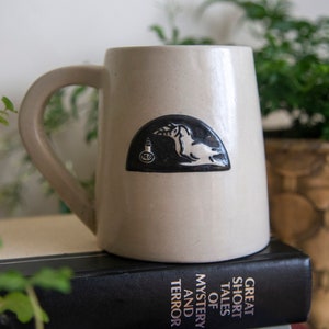 Puede incluir: Una taza de cerámica beige con una ilustración en blanco y negro de una criatura sosteniendo una bombilla. La taza está encima de una pila de libros con el título "Great Short Tales of Mystery and Terror".
