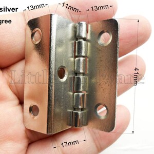 Peut inclure: Une charnière à 90 degrés de couleur argentée, avec une base de 17 mm et une hauteur de 41 mm. La charnière comporte une série de joints cylindriques et trois trous de chaque côté pour le montage. Le texte "color: silver" et "90 degree" sont visibles.