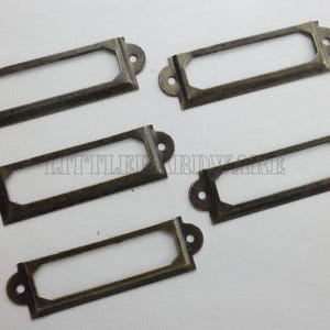 Puede incluir: Cinco portaetiquetas de bronce antiguo. Cada uno tiene un diseño rectangular con bordes redondeados, una abertura central para la etiqueta y dos orificios de montaje. Adecuado para cajones o armarios.