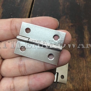 Può includere: Una cerniera in metallo argentato con quattro fori. La cerniera è tenuta in una mano.