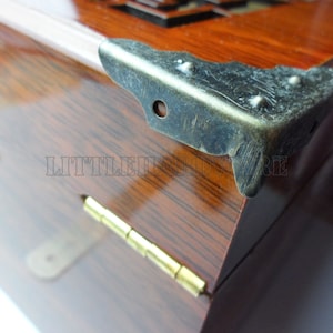 May include: Close-up of a wooden box with a brass hinge and a metal corner protector. The box has a dark wood grain and a glossy finish.