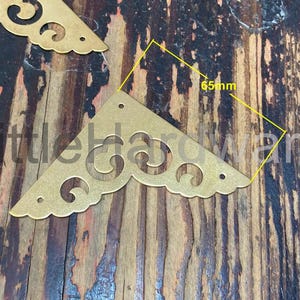 May include: Gold-coloured metal corner brackets with decorative cut-out designs. Each bracket is 6.5cm in length and has a scalloped edge. The brackets are shown on a wooden surface.