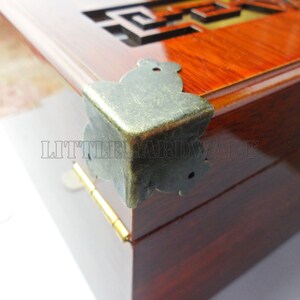 May include: A close-up of a wooden box with a brass corner protector. The box has a dark red finish and a clear glass top. The corner protector is ornate and has a decorative design.