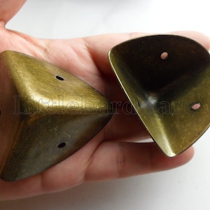 May include: Two antique gold corner protectors with three holes each. The protectors are made of metal and have a distressed finish.