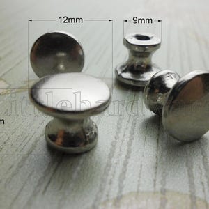 Puede incluir: Tres pomos de metal plateado con una parte superior lisa y redondeada y una base cónica. Las líneas técnicas indican un diámetro de 12 mm para la parte superior, 9 mm para la base y una altura total de 10 mm para estos herrajes.