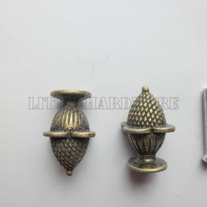 May include: Two antique brass-finished metal cabinet knobs shaped like acorns with textured, cross-hatched detailing on the caps and vertical ridges on the bases, accompanied by a single silver-colored metal mounting screw for installation.