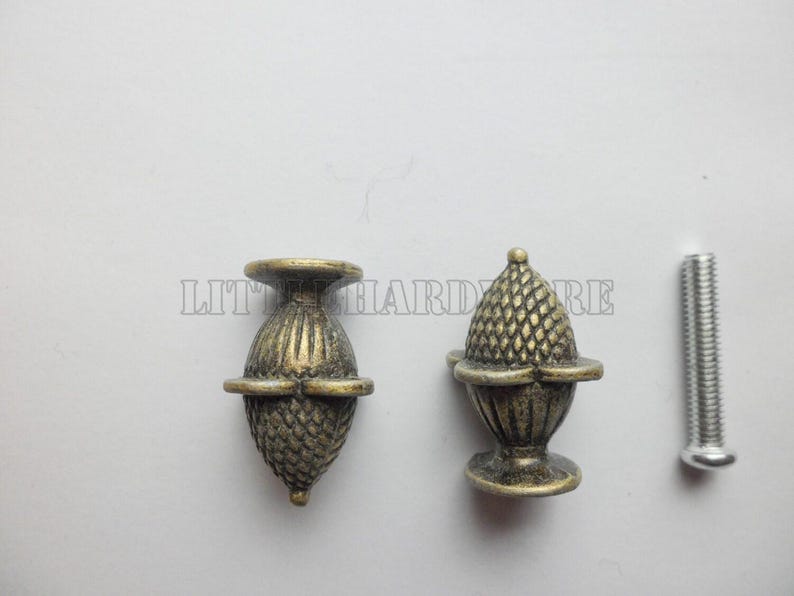 May include: Two antique brass-finished metal cabinet knobs shaped like acorns with textured, cross-hatched detailing on the caps and vertical ridges on the bases, accompanied by a single silver-colored metal mounting screw for installation.