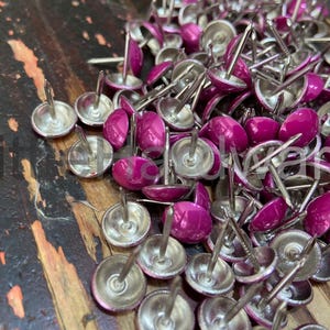 May include: A collection of magenta-headed upholstery tacks with silver bases. The tacks have long pins. The image is a close-up, highlighting the details of the tacks. The tacks are scattered on a wooden surface.