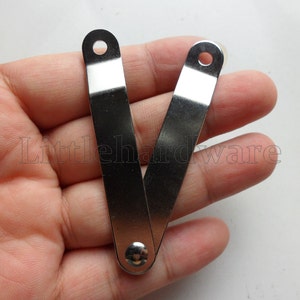 May include: A shiny, silver-coloured metal hinge with two rectangular arms connected by a rivet. Each arm has a small, circular hole at the end. The hinge is open, displaying its folding mechanism. The text "Littlehardware" is partially visible.