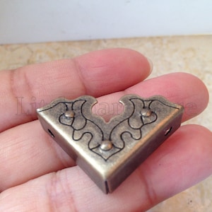 May include: A brass corner bracket with a decorative floral design. The bracket has a triangular shape and is designed to be used for furniture or other projects.