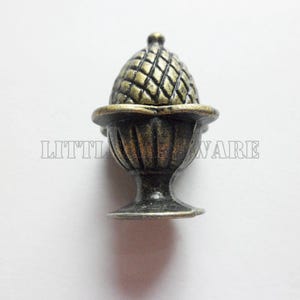 May include: A bronze-toned metal cabinet knob shaped like an acorn resting in a small pedestal cup. The top portion features a cross-hatched texture, while the base has vertical fluting. The text LITTLE HARDWARE is visible in the background.