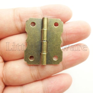 Può includere: Una piccola cerniera in ottone antico con un bordo decorativo. La cerniera ha due piastre rettangolari con angoli arrotondati e tre fori ciascuna per il montaggio. La cerniera è collegata da un perno centrale. Il testo "Little Hardware" è visibile.
