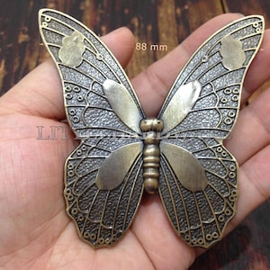 Könnte beinhalten: Ein schmetterlingsförmiges Metallornament mit antikem Messing-Finish, das texturierte Flügel und glatte, erhabene Flügelsegmente aufweist. Das Stück misst 88 mm in der Breite. Der Text "LITTLE HARDWARE" ist in der Mitte des Schmetterlings sichtbar.