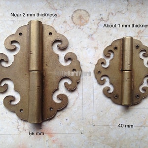 May include: Two antique brass hinges with decorative, floral-like edges. The larger hinge measures 56 mm wide and 60 mm tall, with a thickness of about 2 mm. The smaller hinge is 40 mm wide and 41 mm tall, about 1 mm thick.